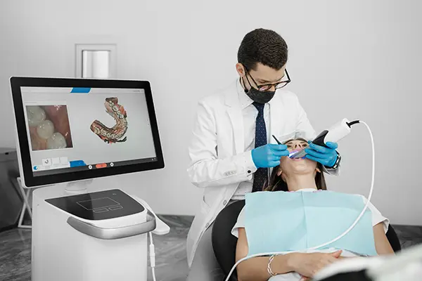 White patient relaxing while her white dentist uses an intra-oral scanner to take a digital impression of her teeth, displayed on a monitor nearby