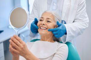 Smiling elderly woman at dental appointment holding mirror, comparing All-on-X and implant-supported dentures
